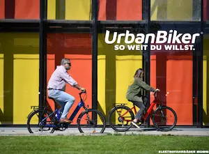 UrbanBiker E-Bikes – Mann und Frau fahren auf zwei E-Bikes auf der Straße vor buntem Gebäude