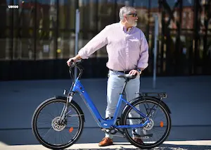 UrbanBiker E-Bikes – Person hält auf einem blauen E-Bike auf der Straße vor dunklem Glasgebäude