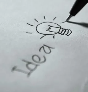 Innovationen: Glühbirne und Kugelschreiber schreiben das Wort ‚Ideen‘ auf grauem Blatt – Symbol für kreative Konzepte und neue Ideen.