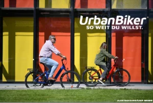 UrbanBiker E-Bikes – Mann und Frau fahren auf zwei E-Bikes auf der Straße vor buntem Gebäude